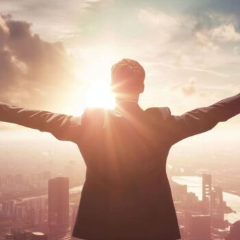 Businessman with arms raised facing sunrise over city skyline expressing success and ambition