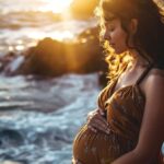 Pregnant woman standing by the sea at sunset, cradling her belly with waves crashing in the background.