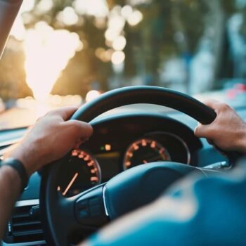 Driver's hands on steering wheel with dashboard view inside car during sunset.