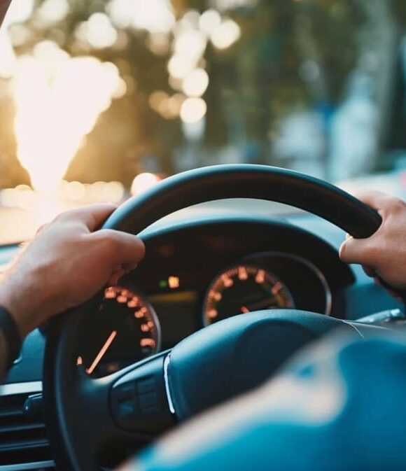 Driver's hands on steering wheel with dashboard view inside car during sunset.